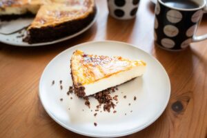 A slice of creamy cheesecake on a plate with coffee mugs on a wooden table.